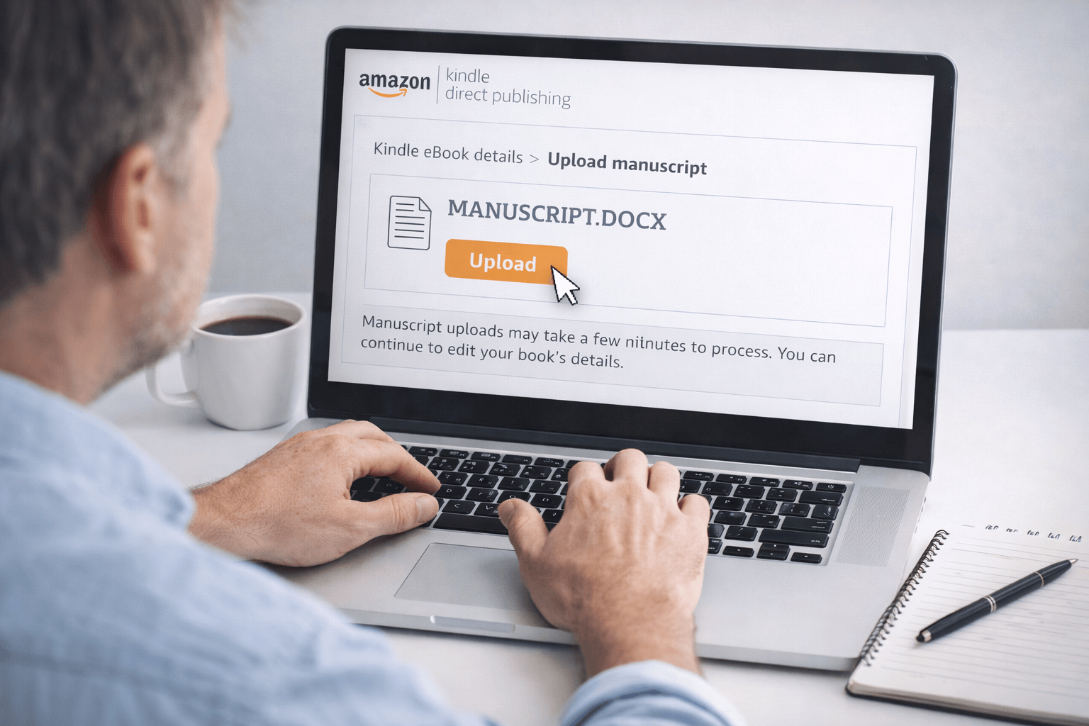 A man is seated at a white desk, uploading a document titled "MANUSCRIPT.DOCX" to Amazon Kindle Direct Publishing (KDP) on his laptop. The screen shows the KDP interface with a large orange “Upload” button and a message below indicating the upload process may take a few minutes. His right hand is near the keyboard, while a white coffee mug sits to the left of the laptop. On the right side of the desk is a lined notepad with a black pen resting on top. The workspace is clean and lit with soft, natural light.
